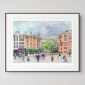 A framed watercolour-style art print depicting Edinburgh Grassmarket with buildings, pedestrians, and a green tree, shown against a light grey wall.