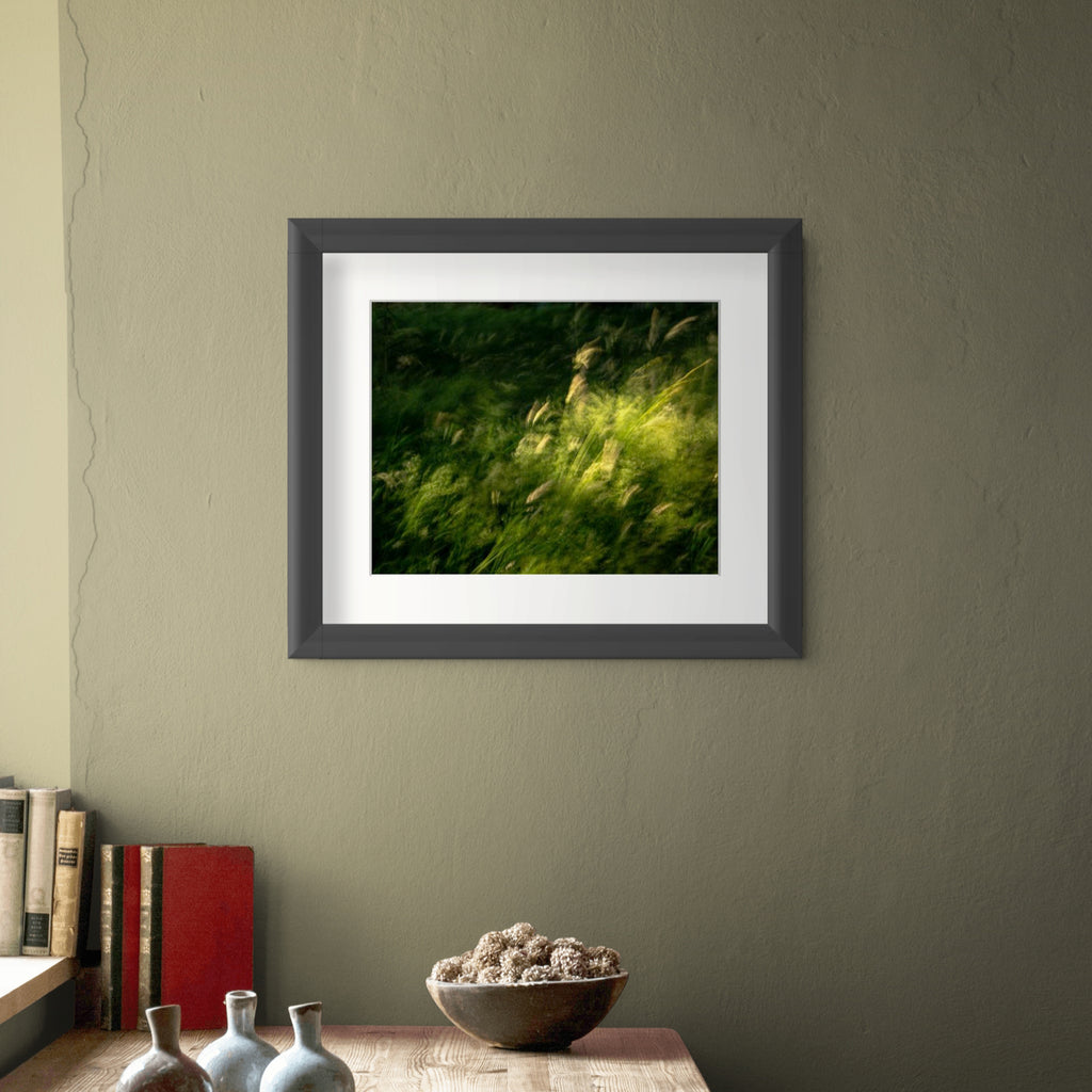 A framed art print featuring a green and yellow grassy landscape swaying in the wind, displayed on a textured olive green wall above a wooden shelf with books and decorative vases.