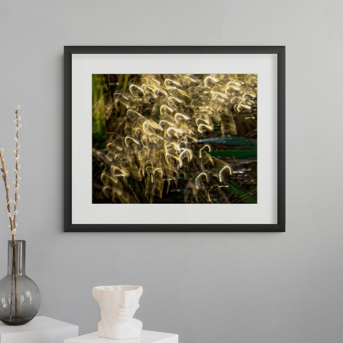 A framed abstract photographic print hangs on a light gray wall above a white side table. The print features golden light trails over dark green foliage, presented in a black frame with a white mat.
