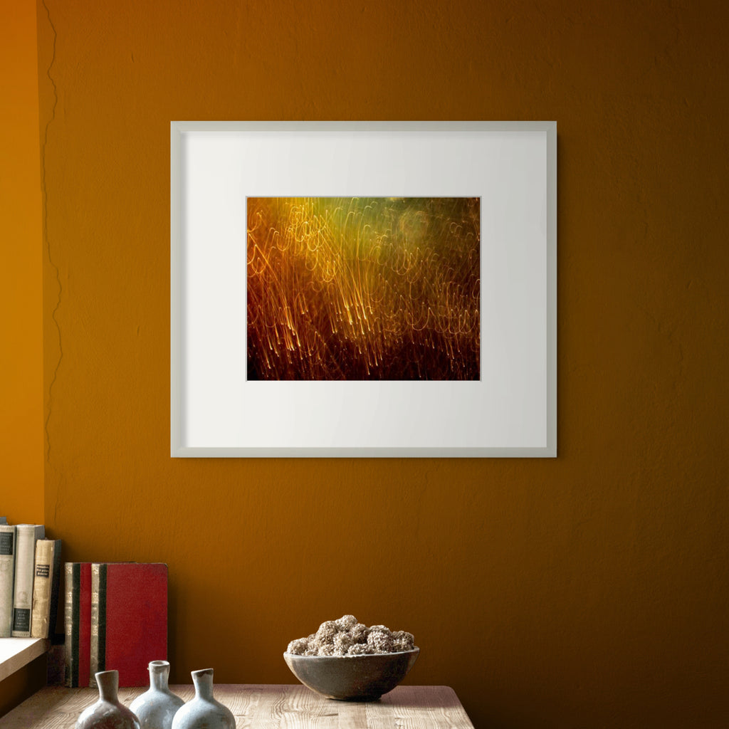 A framed abstract art print featuring golden and brown grass-like textures, displayed on a warm brown wall above a wooden shelf with books and decorative vases.