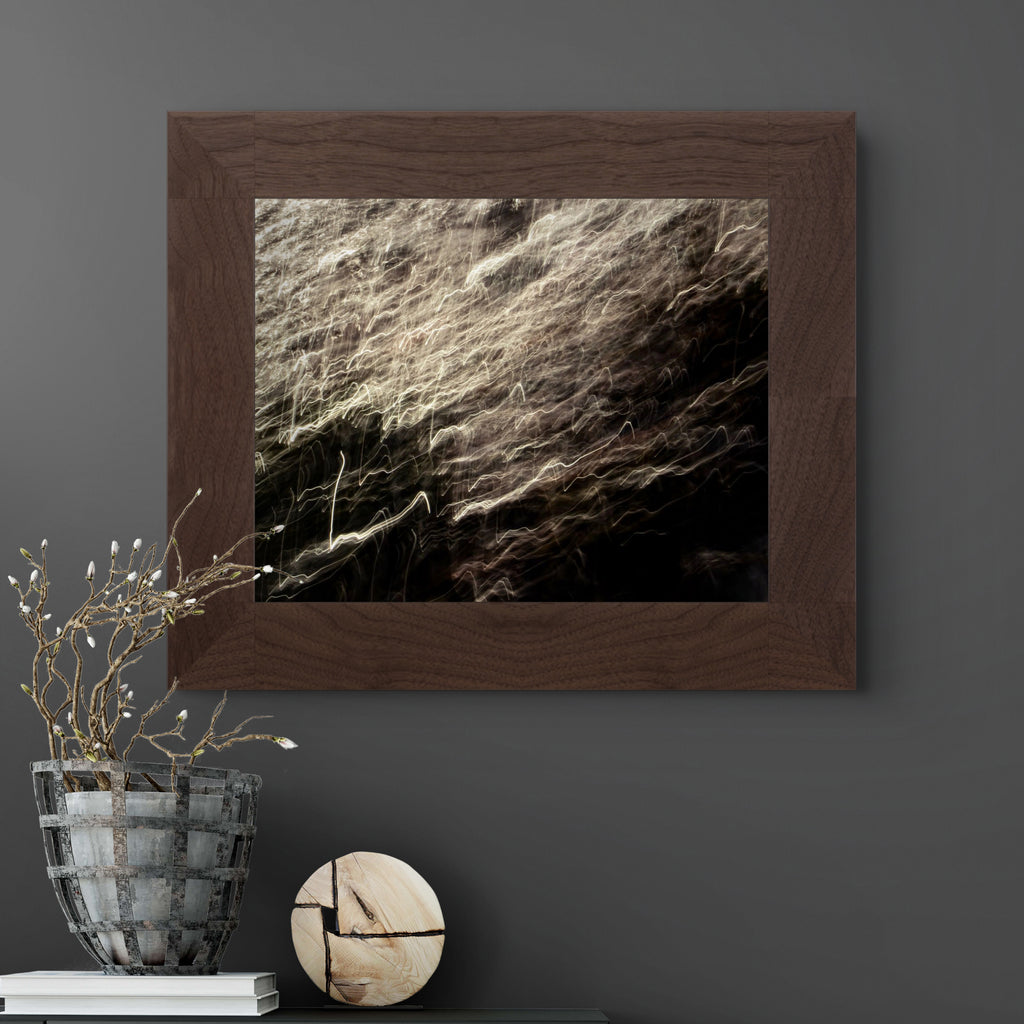 A framed abstract art print featuring textured, flowing light trails in beige, brown, and black, housed in a dark brown wood frame and displayed on a dark grey wall above a table with decorative items.