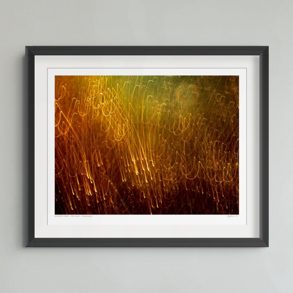 A framed abstract art print featuring glowing golden and orange light trails against a dark brown background, presented in a black frame with a white mat on a light gray wall.