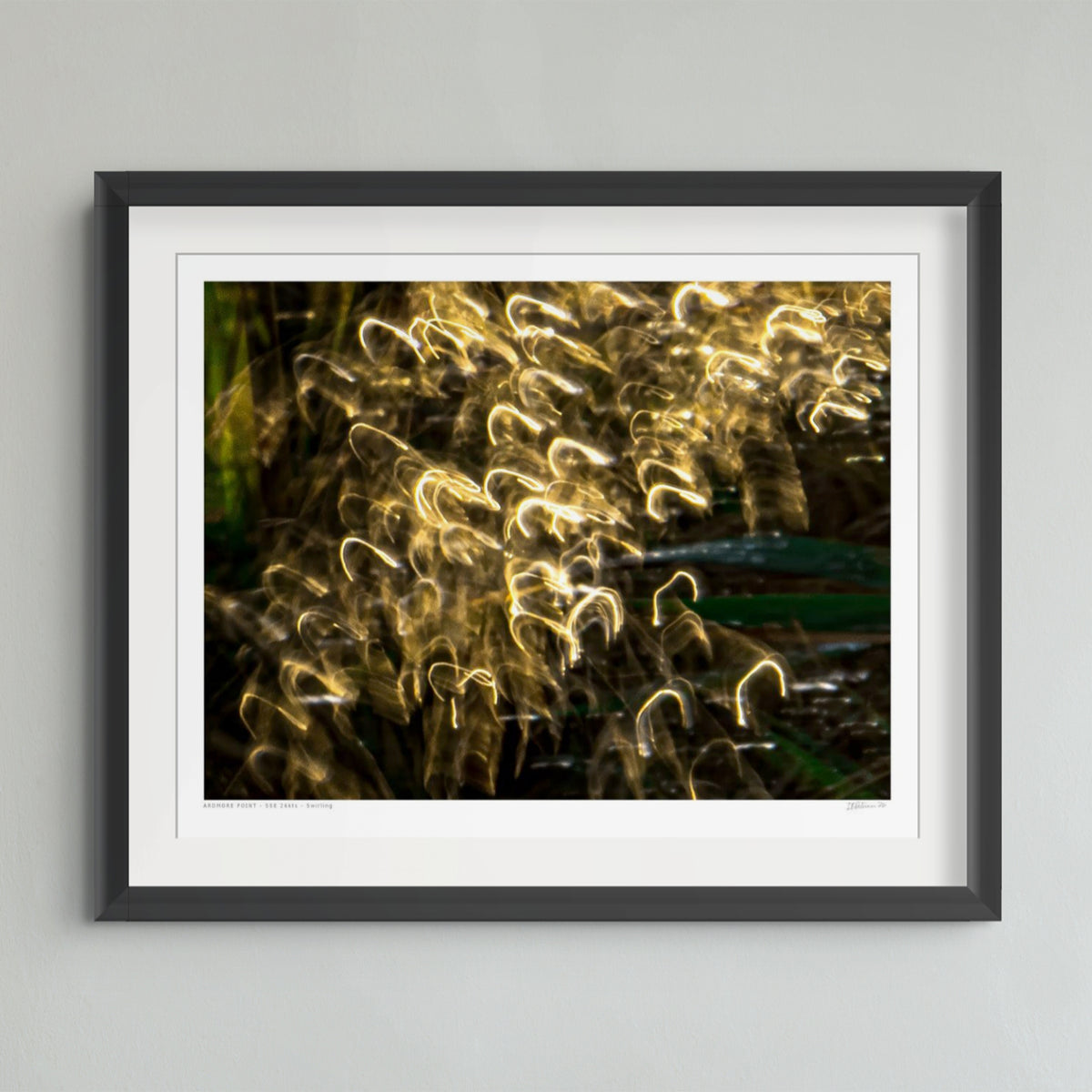A framed abstract photographic print featuring glowing light trails in warm gold and yellow tones against a dark background, presented in a black frame with a white mat on a light gray wall.