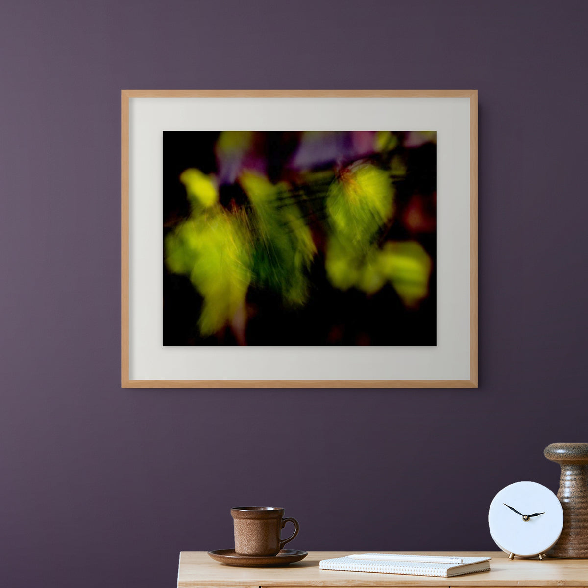 A framed abstract art print featuring vibrant green and yellow bokeh on leaves swaying in the wind, on a black background, styled on a wooden table against a dark purple wall.
