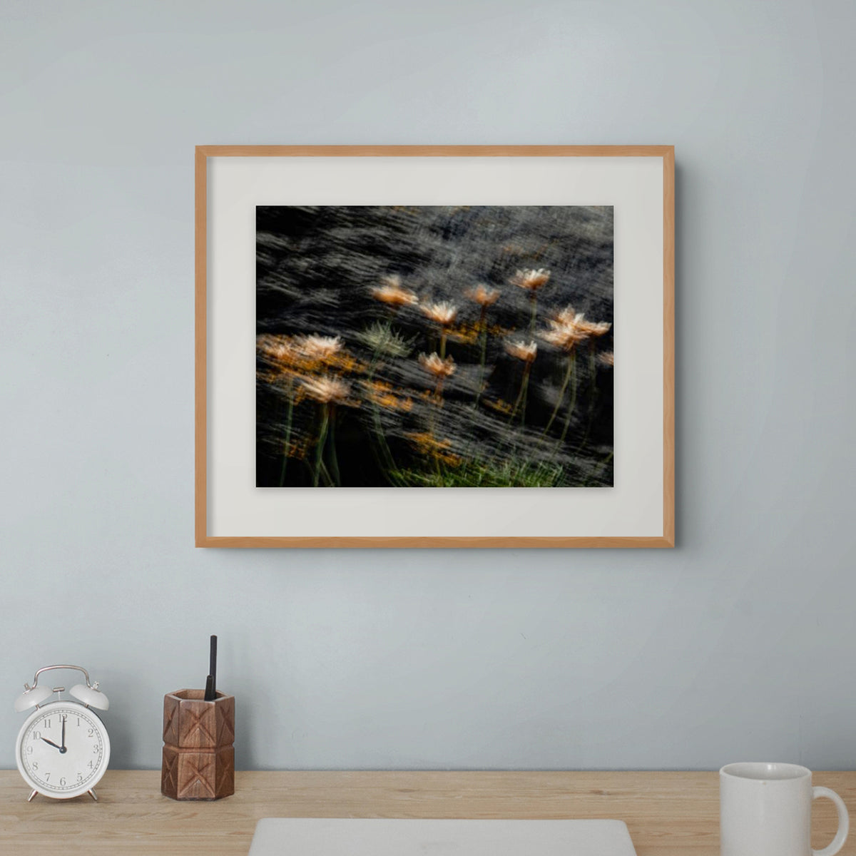 A framed art print depicting a dark, moody scene of wildflowers swaying in the wind against a black background, styled on a light blue-grey wall above a wooden desk with a clock, pen holder, and mug.