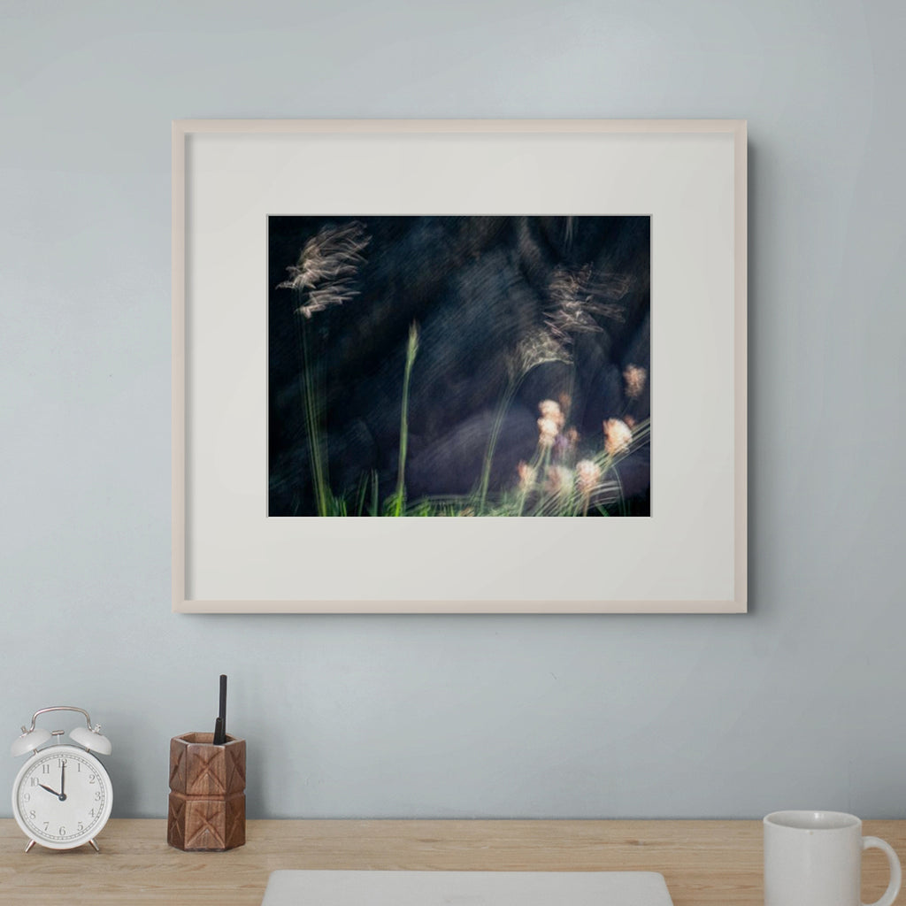 A framed art print featuring a dark, moody photograph of wild grasses and soft pink flowers, displayed on a light blue-gray wall above a wooden desk with a white clock, a geometric pencil holder, and a white mug.