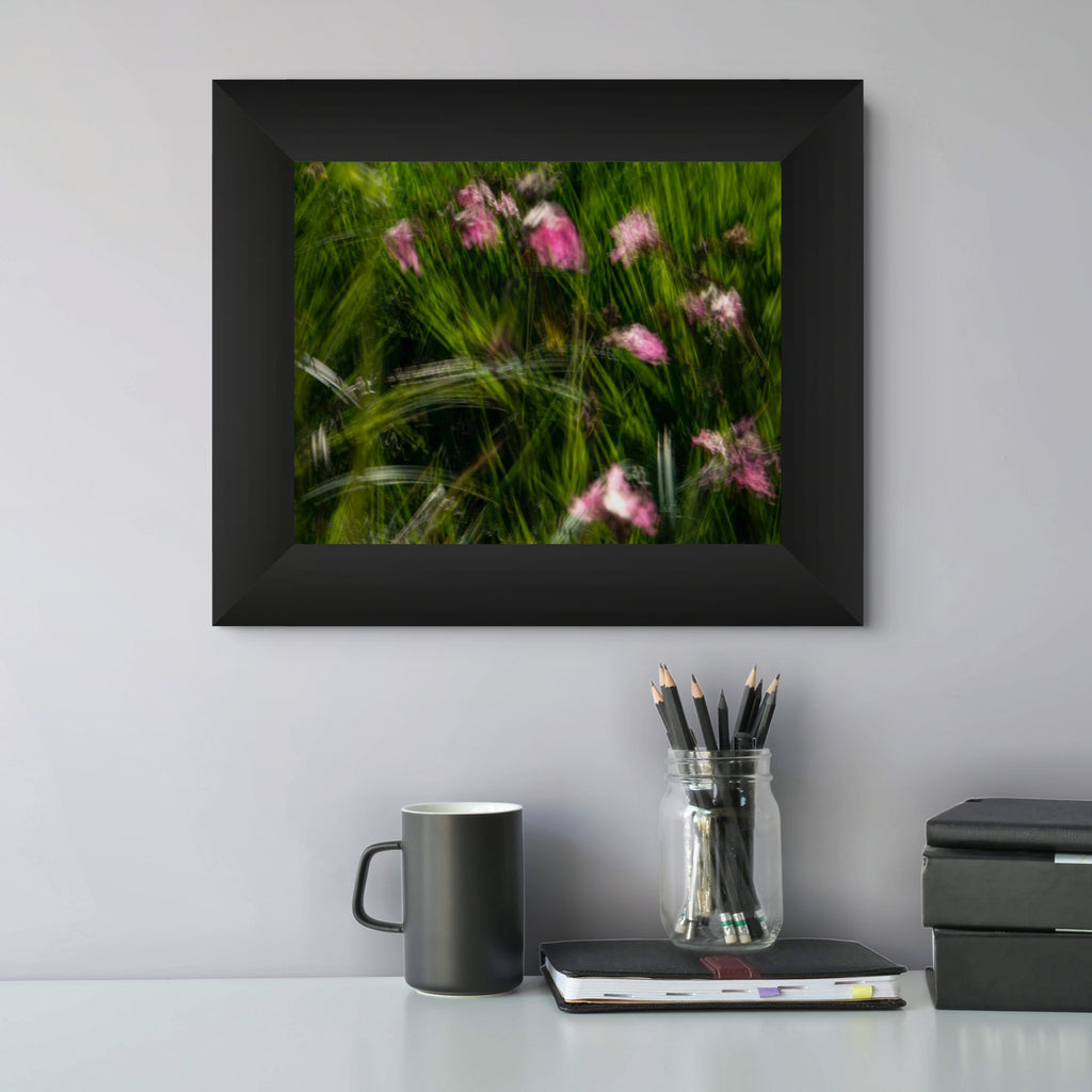 A framed art print depicting green grass with pink wildflowers swaying in the wind, displayed on a light grey wall above a desk with a mug, pencils, and notebooks.
