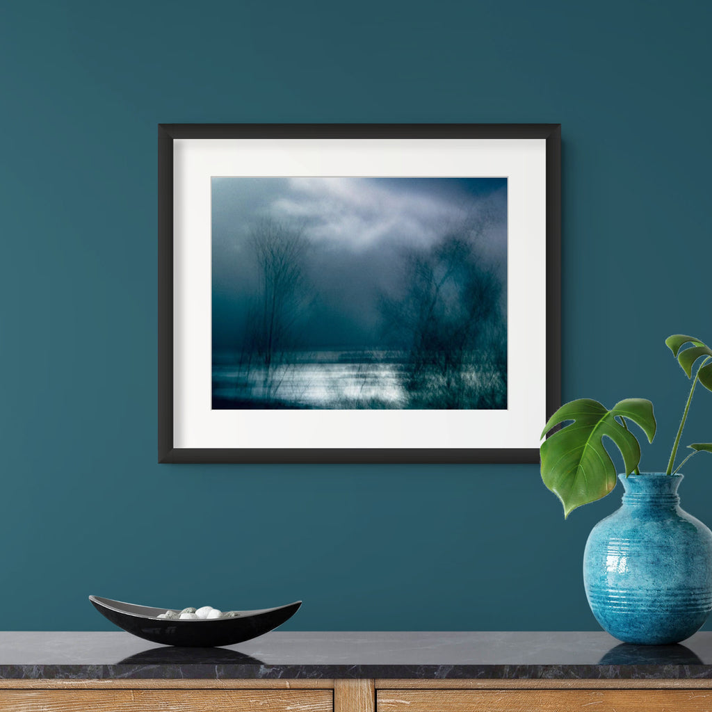 A framed art print featuring a moody, misty landscape in deep teal and black tones, styled on a dark teal wall above a console table with a blue vase and green plant.