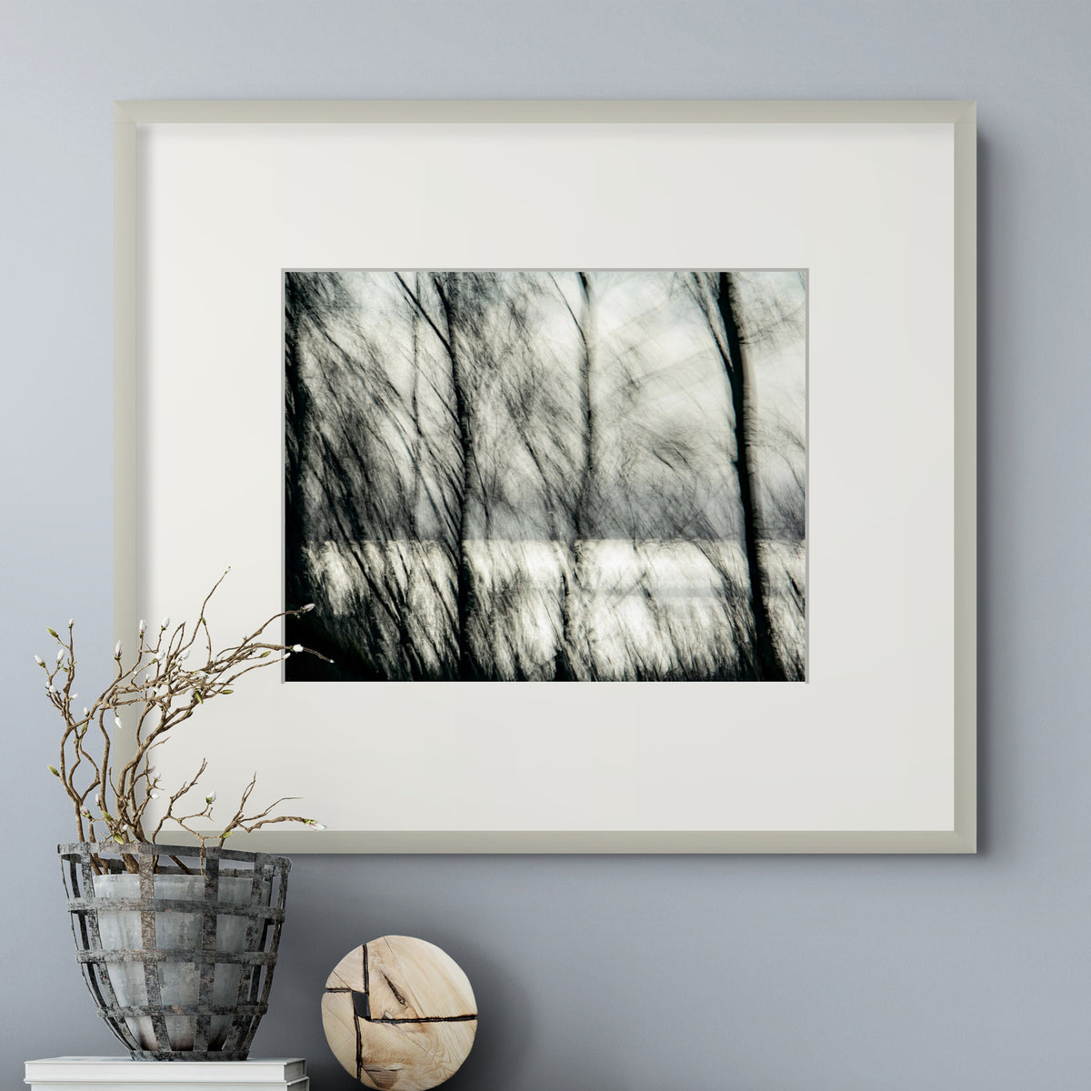 A framed black and white landscape photograph of bare tree branches over a snowy field, presented in a light gray frame with a white mat, styled on a gray wall above a decorative basket and wooden sphere.