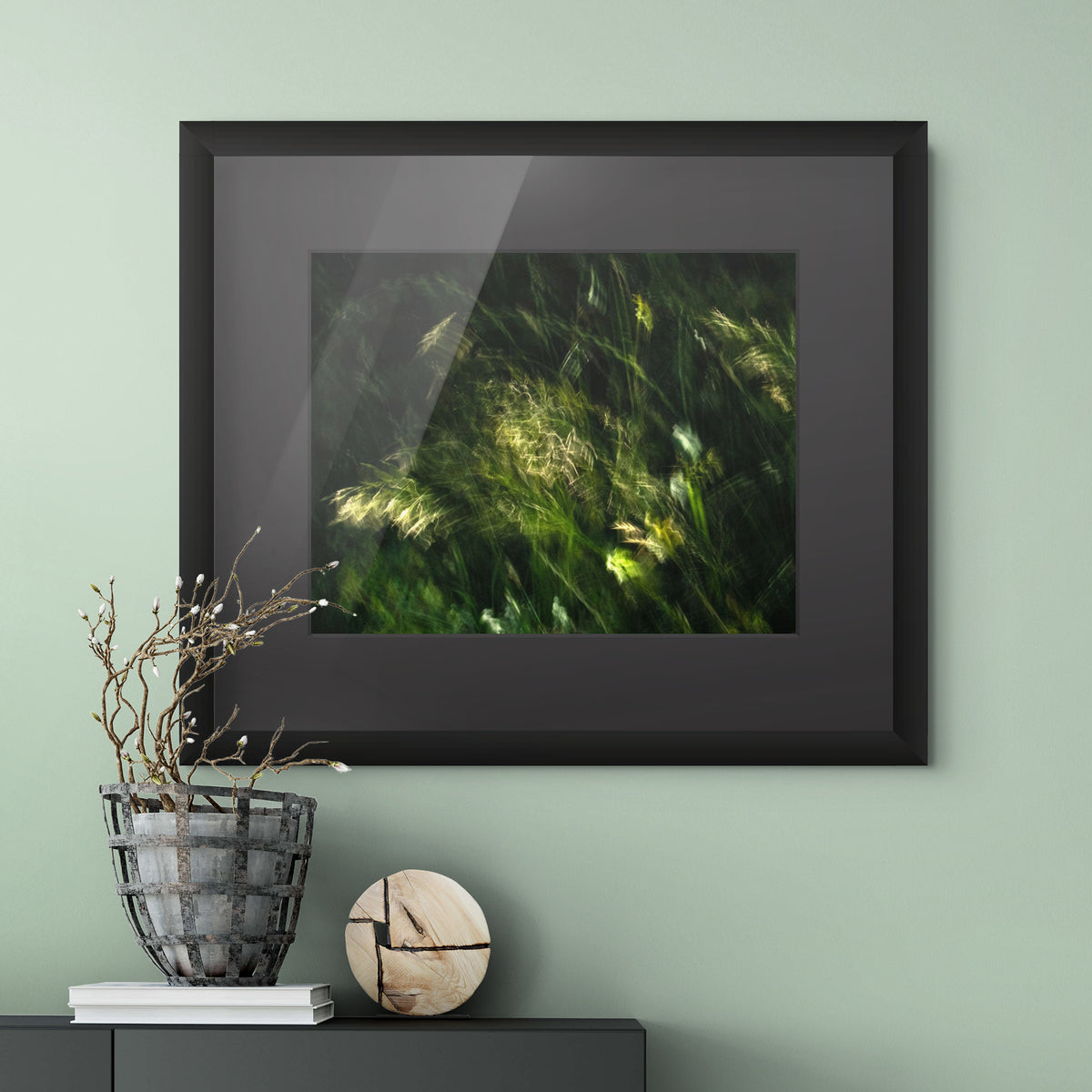 A framed wall art print showing green grasses swaying in the wind with light filtering through, mounted on a pale green wall above a dark console table with a wire basket vase and decorative objects.
