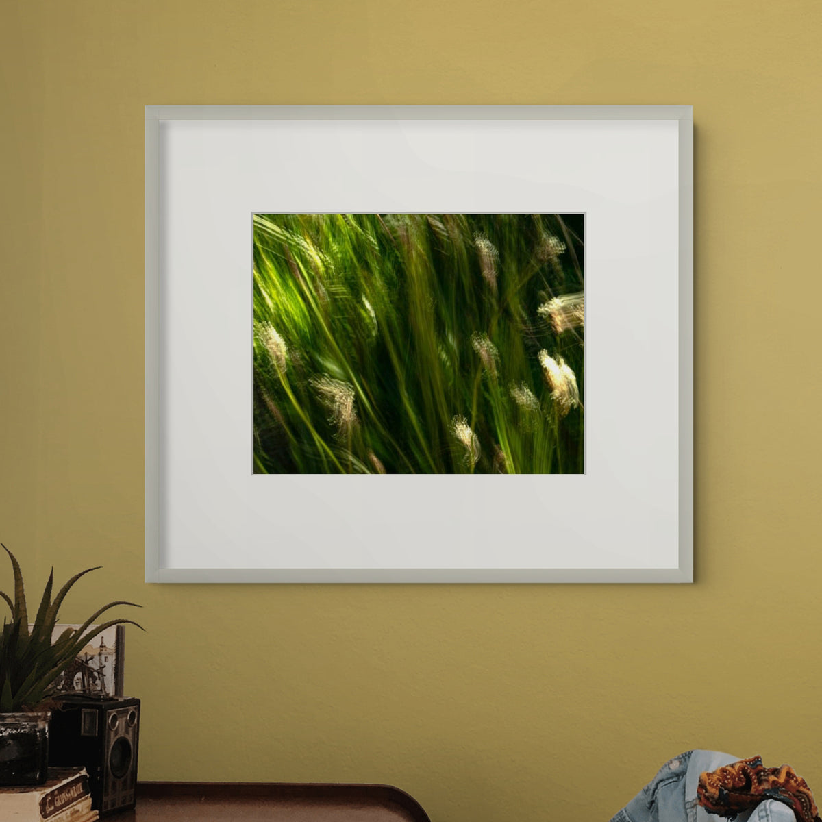 A framed art print features a close-up photograph of green grasses with light-coloured seed heads blowing in the wind, displayed on a mustard-yellow wall above a table with decorative items.